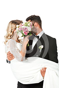 young newlyweds covering faces with bridal bouquet while groom carrying bride