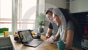 Young nice caucasian couple sitting at table in front of laptop skyping with their happy positive tanned friends from