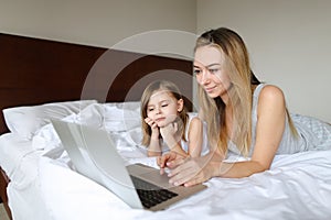 Young nice mother lying in bed with little daughter and using laptop.