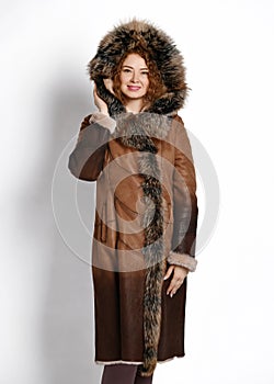 Young nice woman with curly red hair in long brown gradient sheepskin coat with large fluffy fur hood and cuffs on white