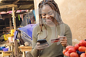 young nigerian woman selling in a local nigerian market using her mobile phone and credit card to do a transaction online smiling