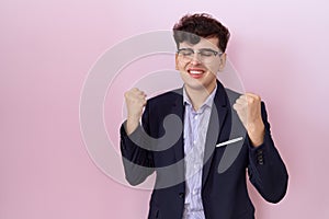 Young non binary man with beard wearing suit and tie very happy and excited doing winner gesture with arms raised, smiling and