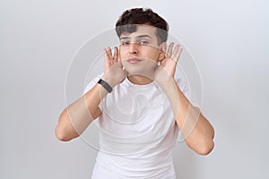 Young non binary man wearing casual white t shirt trying to hear both hands on ear gesture, curious for gossip