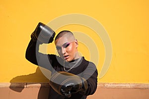 Young non-binary person wears boxing gloves on a mustard yellow background, the person is make up and does different boxing poses