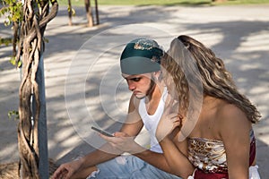 Young, non-conformist Latino and Hispanic boy and girl couple sitting on a bench checking social networks on their cell phone.