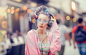 Young nonconformist girl listening music in headphones on the crowded streets. Blurred urban background. Vanguard fashion.