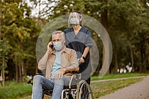 Young nurse in face shield and protective face mask taking care of senior handicapped patient in wheelchair talking on