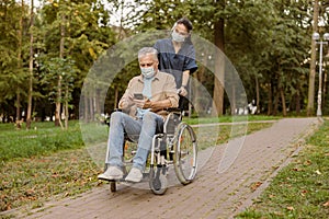 Young nurse in face shield and protective face mask taking care of senior handicapped patient in wheelchair using