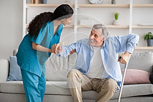 Young nurse helping elderly man with crutches to get up from sofa indoors