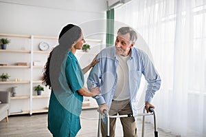 Young nurse helping older man to walk with frame at retirement home. Professional eldercare service