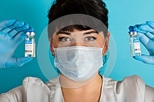 Young nurse in a medical mask holding two vials of the Covid-19 coronavirus vaccine on a blue background. Close-up