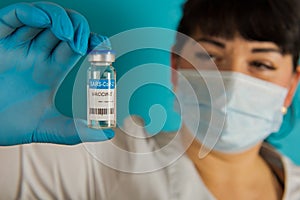 Young nurse in a medical mask holding vial of the Covid-19 coronavirus vaccine on a blue background. Close-up. Selective focus
