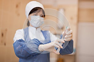 Young nurse in special sterile clothing in a protective mask is standing indoors holding a syringe in her hand.