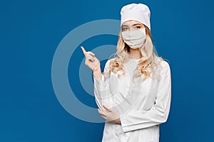 A young nurse in a white coat and medical mask poses on a blue background and points a finger to the left side, isolated