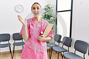 Young nurse woman at medical clinic waiting room celebrating victory with happy smile and winner expression with raised hands