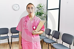 Young nurse woman at medical clinic waiting room scared and amazed with open mouth for surprise, disbelief face