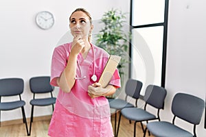 Young nurse woman at medical clinic waiting room serious face thinking about question with hand on chin, thoughtful about