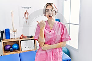 Young nurse woman working at pain recovery clinic smiling with happy face looking and pointing to the side with thumb up