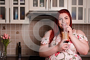 Young obese woman smiling while drinking a shake in the kitchen. Bodypositive and improper nutrition.