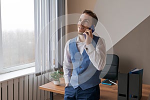 Young office worker is talking smiling on phone leaning against desktop.