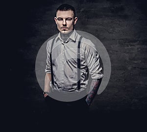 Young old-fashioned tattooed guy wearing white shirt and suspenders, looking at a camera on a dark background.