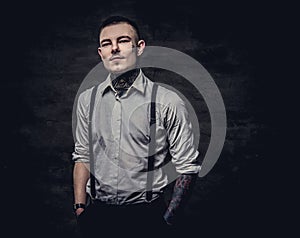 Young old-fashioned tattooed guy wearing white shirt and suspenders, looking at a camera on a dark background.