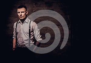 Young old-fashioned tattooed guy wearing white shirt and suspenders, looking at a camera on a dark background.