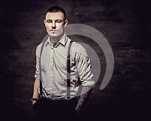 Young old-fashioned tattooed guy wearing white shirt and suspenders, looking at a camera on a dark background.