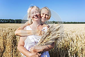 Young and old women in white sundresses in a wheat field on a sunny day, holding ears of corn in their hands, smiling and hugging