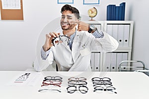 Young optician man holding optometry glasses pointing with hand finger to face and nose, smiling cheerful