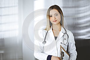 Young optimistic woman-doctor is holding a clipboard in her hands, while standing in a clinic. Portrait of friendly