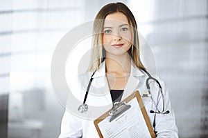 Young optimistic woman-doctor is holding a clipboard in her hands, while standing in a clinic. Portrait of friendly