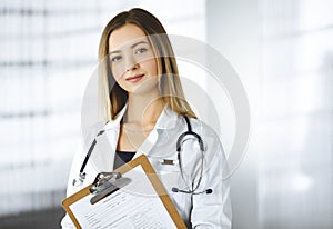 Young optimistic woman-doctor is holding a clipboard in her hands, while standing in a clinic. Portrait of friendly