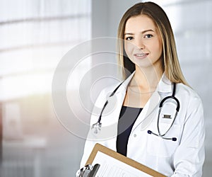 Young optimistic woman-doctor is holding a clipboard in her hands, while standing in a sunny clinic. Portrait of