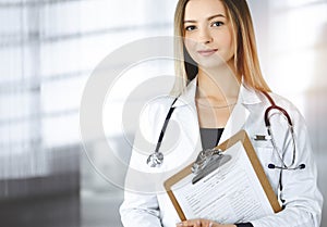 Young optimistic woman-doctor is holding a clipboard in her hands, while standing in a sunny clinic. Portrait of
