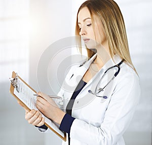 Young optimistic woman-doctor is holding a clipboard in her hands, while standing in a sunny clinic. Portrait of
