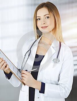 Young optimistic woman-doctor is holding a clipboard in her hands, while standing in a sunny clinic. Portrait of