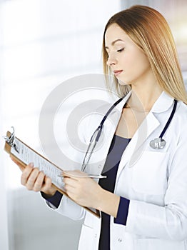 Young optimistic woman-doctor is holding a clipboard in her hands, while standing in a sunny clinic. Portrait of