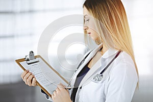 Young optimistic woman-doctor is holding a clipboard in her hands, while standing in a sunny clinic. Portrait of