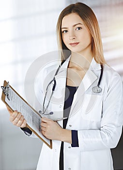 Young optimistic woman-doctor is holding a clipboard in her hands, while standing in a sunny clinic. Portrait of