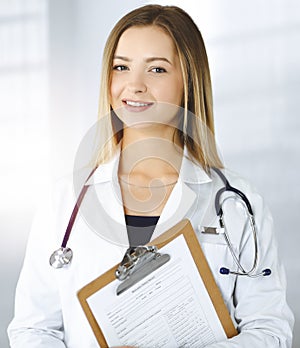 Young optimistic woman-doctor is holding a clipboard in her hands, while standing in a sunny clinic. Portrait of