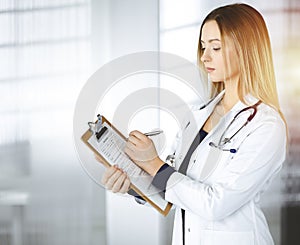 Young optimistic woman-doctor is holding a clipboard in her hands, while standing in a sunny clinic. Portrait of