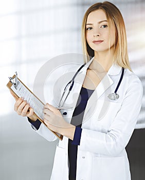 Young optimistic woman-doctor is holding a clipboard in her hands, while standing in a sunny clinic. Portrait of