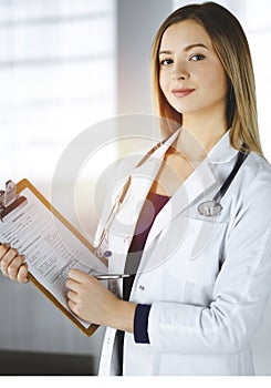 Young optimistic woman-doctor is holding a clipboard in her hands, while standing in a sunny clinic. Portrait of