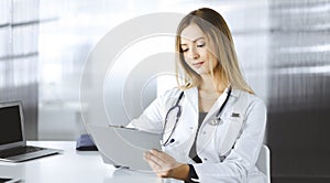 Young optimistic woman-doctor is using a clipboard, while sitting at the desk in her cabinet in a clinic. Portrait of