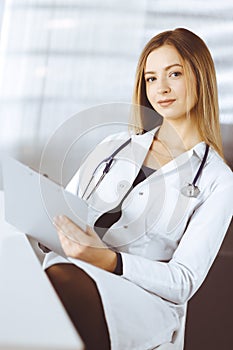 Young optimistic woman-doctor is using a clipboard, while sitting at the desk in her cabinet in a clinic. Portrait of