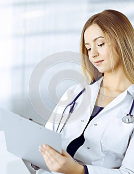 Young optimistic woman-doctor is using a clipboard, while sitting at the desk in her cabinet in a clinic. Portrait of