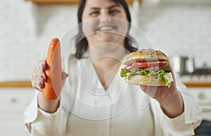 Young overweight plus size fat woman holding a burger in one hand and a carrot in other.