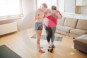 Young overweight and slim women in living room. Plus size model stand on weight scale. They look down. Daylight. Body