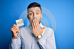 Young overworked business man asking for help holding paper over blue background cover mouth with hand shocked with shame for